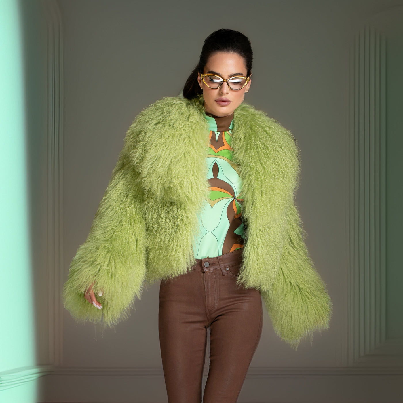 Person wearing a bright green fur coat and brown pants in a room with teal walls.