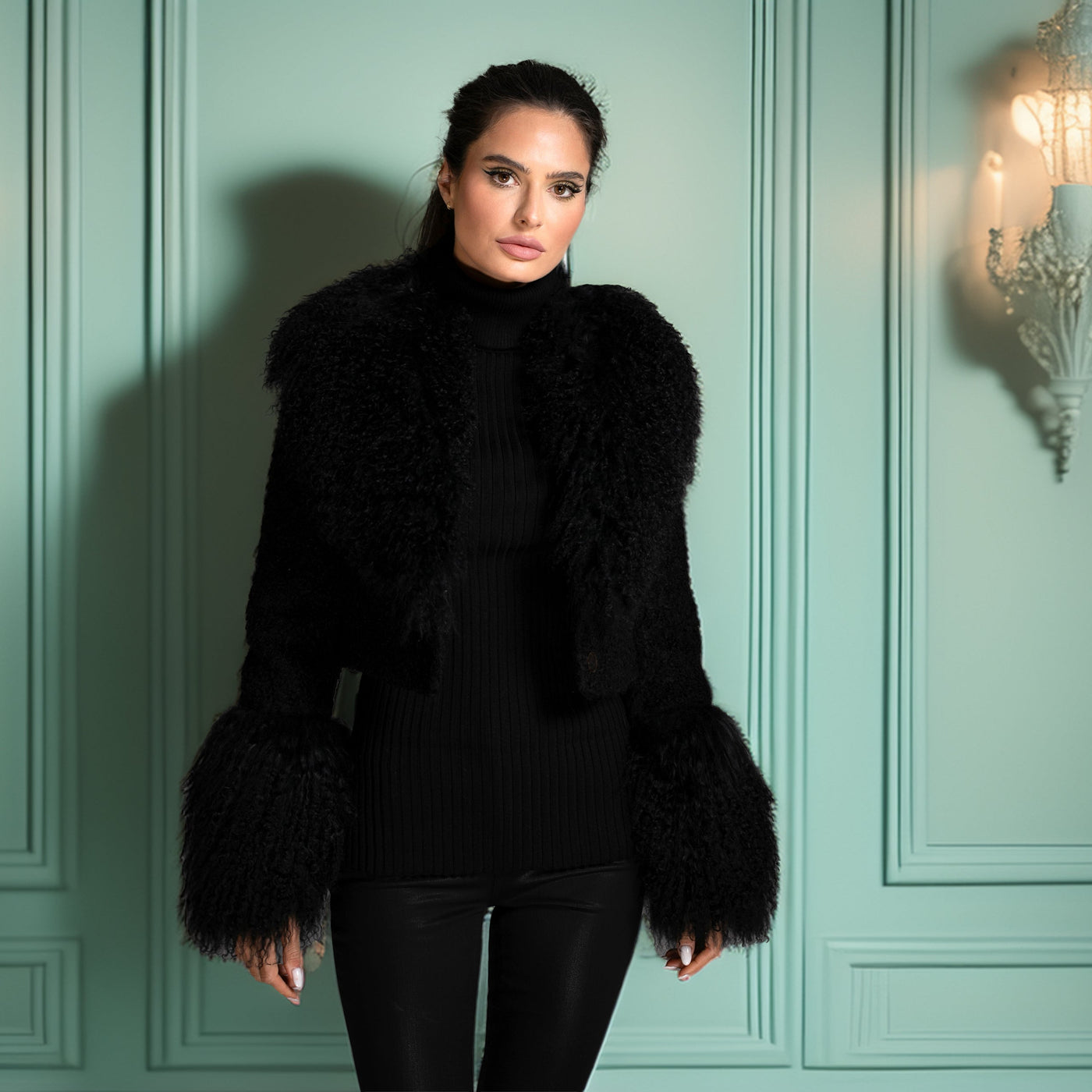 Woman wearing a black fur coat against a teal paneled wall with chandelier lights.